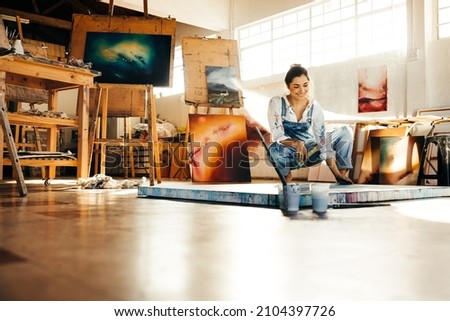 Happy female painter making a blue painting on a canvas. Creative young woman smiling while looking at her painting on the floor. Cheerful young artist working on a new project in her atelier. Royalty-Free Stock Photo #2104397726
