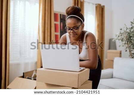 Happy beautiful girl leans over laptop standing on cardboard boxes, browses online stores, orders furniture for new apartment she moved into