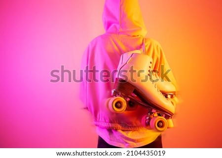 Teenager girl in hooded sweatshirt carrying classic leather roller skates. Pink and orange background, pop art style. Sports equipment and recreation. Layout with free copy (text) space.