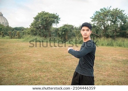 A young asian man doing standing oblique twists. In preparation for a morning run at an outdoor field. Royalty-Free Stock Photo #2104446359