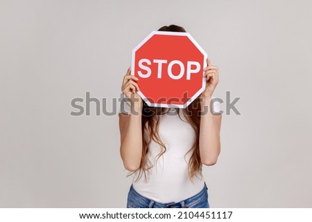 Unknown woman covering face with stop symbol, holding red traffic sign stop, warning of danger, restriction and limits, wearing white T-shirt. Indoor studio shot isolated on gray background.