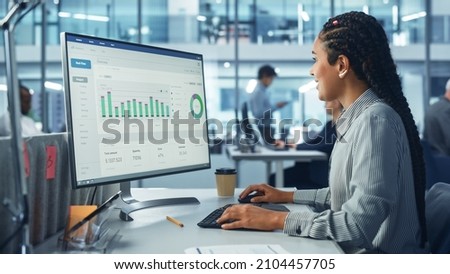 Corporate Office: Black Female IT Technician Using Desktop Computer with Big Data Statistics, Graphs. Smiling Creative Software Engineer Work on eCommerce Project Marketing, Development. Royalty-Free Stock Photo #2104457705