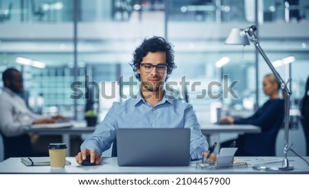 Modern Office Businessman Working on Computer. Portrait of Successful Latin IT Software Engineer Working on a Laptop at his Desk. Diverse Workplace with Professionals. Front View Shot Royalty-Free Stock Photo #2104457900