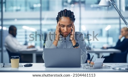 Young Black Woman Office Worker Uses Laptop, Feels Sudden Burst of Pain, Headache, Migraine. Overworked Accountant Feeling Project Pressure, Stress, Massages Her Head, Temples. Front View Portrait Royalty-Free Stock Photo #2104461113
