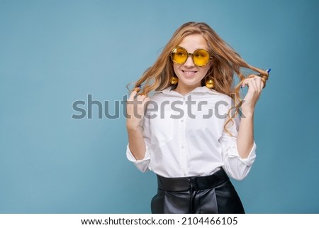 Close up portrait of crazy funny cheerful comic attractive beautiful girl in white shirt cheerfully posing in yellow glasses isolated on blue background. Caucasian teenager grimacing in studio