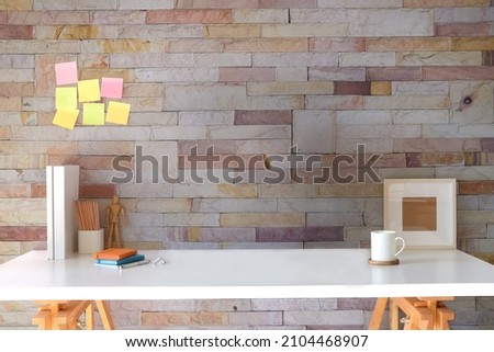 Designer workplace, pencil holder, books, picture frame and coffee cup on white table with brick wall.