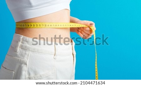 Slim woman measures her waistline with measuring tape. Healthy body shaping weight loss concept. Slim waist small belly in big white denim pants isolated over blue color background. Long web banner Royalty-Free Stock Photo #2104471634