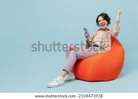 Full body young smiling cheerful cool happy woman 20s wear brown shirt sit in bag chair hold use mobile cell phone do winner gesture isolated on pastel plain light blue background studio portrait.