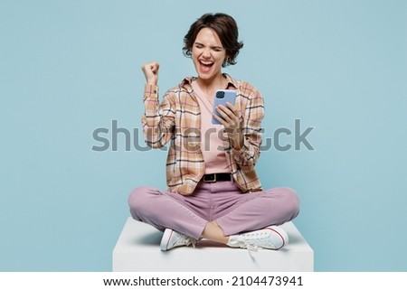 Full body young smiling happy woman 20s wear casual brown shirt sit on white chair use mobile cell phone do winner gesture isolated on pastel plain light blue color background People lifestyle concept Royalty-Free Stock Photo #2104473941