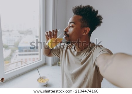 Close up young profile african american man in casual clothes drink orange citrus juice do selfie shot pov mobile phone prepare cooking food in light kitchen at home alone indoor Healthy diet concept.