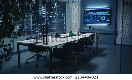 Modern Empty Meeting Room with Big Conference Table with Various Documents and Laptop on it. Wall TV Showing Company's Growth, Statistics, Graphs and Pie Charts. Late Evening or Night City Outside. Royalty-Free Stock Photo #2104484021