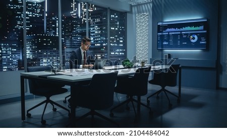 Successful Handsome Businessman Working on Laptop Computer in Big City Office Late in the Evening. Finance Investment Analyst Checking Line and Pie Graphs from Project Management Report. Royalty-Free Stock Photo #2104484042