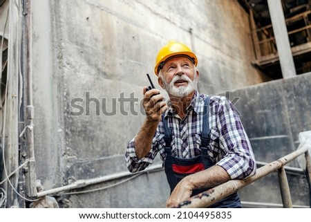 An old factory worker with a helmet on his head, leans on the railing inside of the factory and communicates with his coworkers over the walkie-talkie. Workers talking with walkie-talkie. Royalty-Free Stock Photo #2104495835