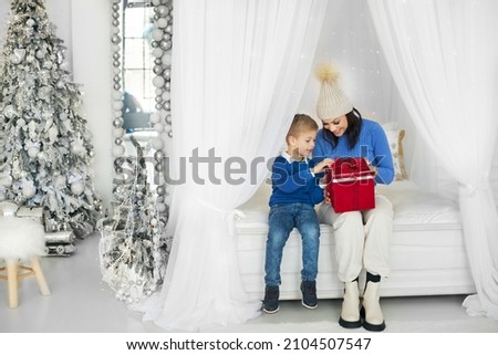 A happy mother is having fun with her son, who opens a wrapped present at home during the celebration. Mother and son are sitting on a white bed and opening a red gift box with a Christmas present.