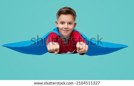 Happy little boy in superhero costume and cape smiling at camera while pretending to fly with outstretched fists against turquoise background