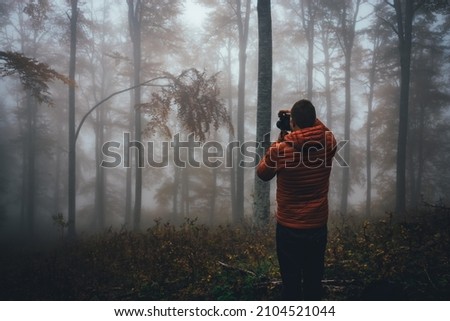 Young bearded hiker wearing orange jacket taking photos at forest with fog and mist. Back view of photographer in field enjoying the view.