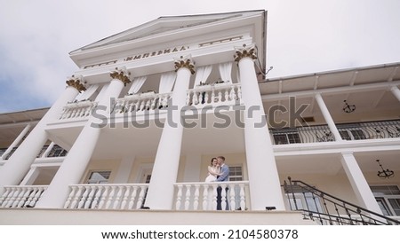 Newlyweds kiss in building with columns. Action. View below is of beautiful white building with columns and newlyweds. Newlyweds in beautiful white building Royalty-Free Stock Photo #2104580378