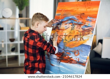 Inspired caucasian boy creating his masterpiece on easel using paints and hands. Little student enjoying art lesson at modern school.
