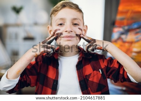 Portrait of pretty caucasian boy painting his cheeks with hands while posing on camera at art studio. Little artist after creative process. Talent and inspiration concept.