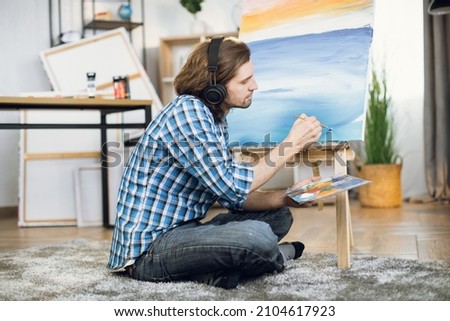 Happy caucasian man sitting on floor in wireless headset and painting with brush and easel. Inspired artist listening favorite music while creating masterpiece in studio.