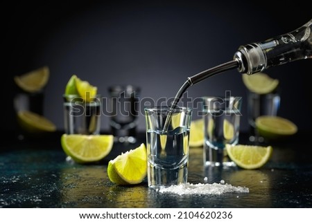 Tequila is poured into a glass. Tequila shots with lime slices and sea salt on a dark blue table. Royalty-Free Stock Photo #2104620236