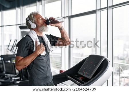 Active senior in grey t-shirt with white towel on shoulders, drinking water from bottle, standing on treadmill. Aged man with headphones staying hydrated after sport session.