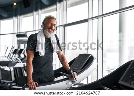 Close portrait happy grey-haired bearded mature man with towel on shoulder, smiling, looking at camera. Handsome sportsman in headphones posing in gym, feeling satisfied after workout. Royalty-Free Stock Photo #2104624787