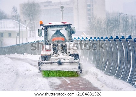 Tractor sweep snow with rotating brush and snowplow from pedestrian zone on bridge, sidewalk snow management during blizzard. Snow plow service remove snow and ice from road, winter road maintenance Royalty-Free Stock Photo #2104681526
