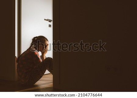 A little girl huddled in a corner and cries, covering her eyes and ears with her hands, because her parents are quarreling. Family problem, violation of children's rights Royalty-Free Stock Photo #2104776644