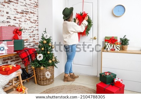 Young fashionable woman adjusting red bow on white door for Christmas in light room