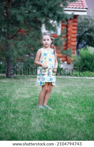 Portrait of little girl looking at camera in surprise with hands near belly having fun wearing light summer dress and slippers in park in daytime. Amazing background full of green. 