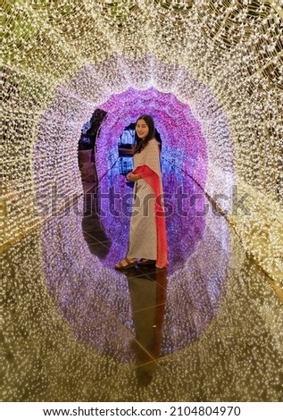 Asian woman in Thai traditional costume standing with colorfully illuminated lighting tunnel with reflection on ground
