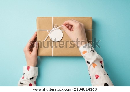 First person top view photo of saint valentine's day decorations female hands in white shirt untying small bow on craft paper giftbox with pricetag on isolated pastel blue background with copyspace
