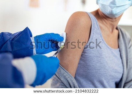 Senior woman patient sitting in a medical clinic and is being given the Covid 19 vaccine in his shoulder by a female doctor, both wearing protective face masks