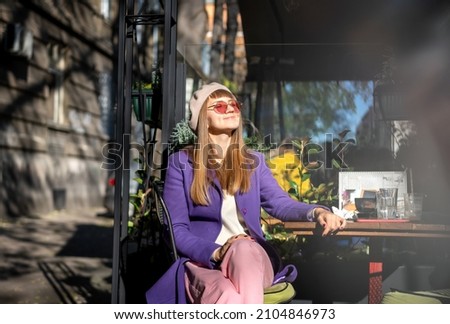 Young cheerful woman in purple coat sitting in street cafe enjoying sunny spring day Royalty-Free Stock Photo #2104846973