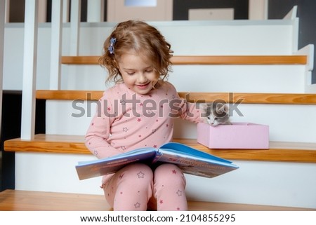 a little girl in pajamas is sitting at home on the stairs reading a book for a kitten