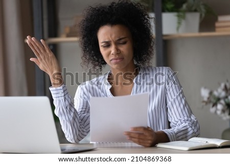 Stressed young African American businesswoman looking at paper document in hands, feeling dissatisfied with bad research results, finding mistakes in financial report, confused with letter. Royalty-Free Stock Photo #2104869368