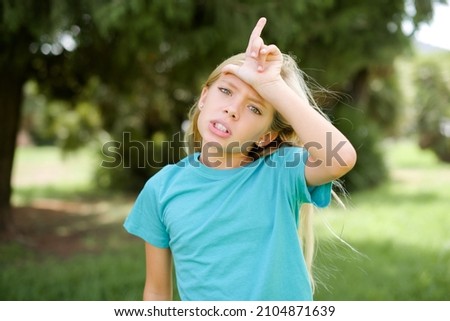 caucasian little girl wearing blue t-shirt standing outdoors making fun of people with fingers on forehead doing loser gesture mocking and insulting.