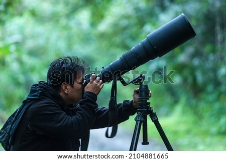 A professional wildlife photographer takes pictures with a digital camera and super-telephoto lens on a tripod in a jungle. Focus on the lens. Royalty-Free Stock Photo #2104881665