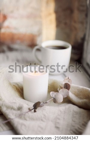 White burning candle, woolen scarf or plaid and cup of hot tea on windowsill inside old house, selective focus. Creating cozy and inviting atmosphere by adding candles to home decor Royalty-Free Stock Photo #2104884347