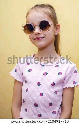 Baby girl in sunglasses on a yellow background. Portrait of a little girl in a vertical photo with round sunglasses. Cute baby posing in a t-shirt with a picture of berries