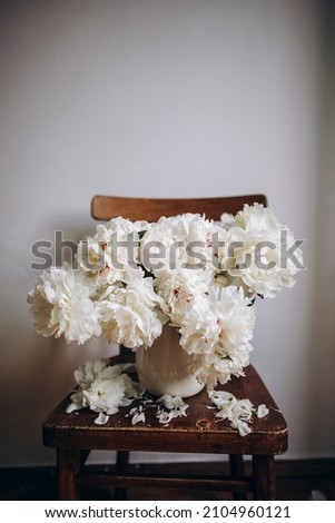 white ceramic vase full of flowers of peonies. a bouquet of white flowers on a wooden chair. fallen white petals. copy the space at the top.