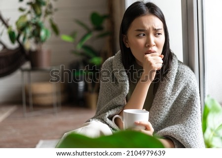 Sad ill asian girl staying on self quarantine during covid, catching flu and sitting at home with cup of tea near window, coughing in hand Royalty-Free Stock Photo #2104979639
