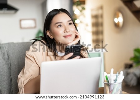 Smiling asian girl with laptop and phone, looking up and thinking, dreaming of something. Woman relaxing at home with computer Royalty-Free Stock Photo #2104991123