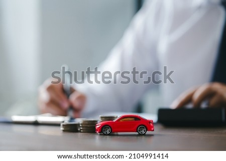 Car on desk with stack coins and man signing purchase documents in background. Red modern car while hand completes the insurance policy or rental documents. Guy buying new car at dealership.