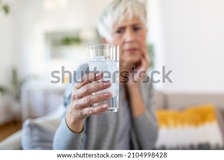 Senior woman with sensitive teeth and hand holding glass of cold water with ice. Healthcare concept. Mature woman drinking cold drink, glass full of ice cubes and feels toothache, pain