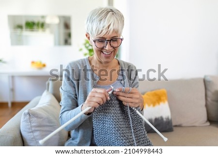 Elderly woman in glasses sit on couch at home smile holding knitting needles and yarn knits clothes for loved ones, favorite activity and pastime, retired tranquil carefree life concept Royalty-Free Stock Photo #2104998443