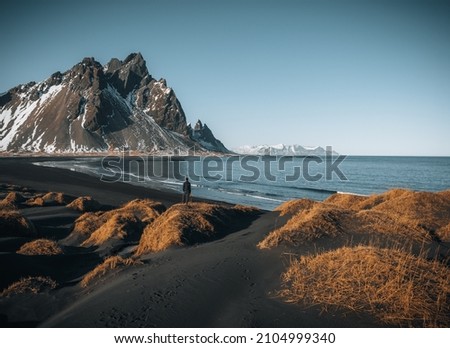 Young man tourist standing at beach during sunset runrise and gorgeous reflection of Vestrahorn mountain on Stokksnes cape in Iceland. Beautiful snow covered mountains. Location: Stokksnes cape Royalty-Free Stock Photo #2104999340