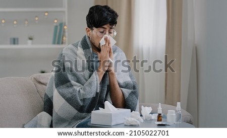 Arab bearded sick sad man with glasses sits on couch at home suffers from runny nose flu disease coronavirus pandemic covid epidemic sneezes. Unwell guy feeling bad fever virus illness symptoms indoor Royalty-Free Stock Photo #2105022701