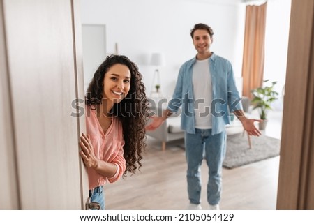 Portrait of cheerful couple inviting guests to enter home, happy people standing in doorway of modern flat, looking out waiting for visitor to come in, receiving friend, lady peeking out of front door Royalty-Free Stock Photo #2105054429